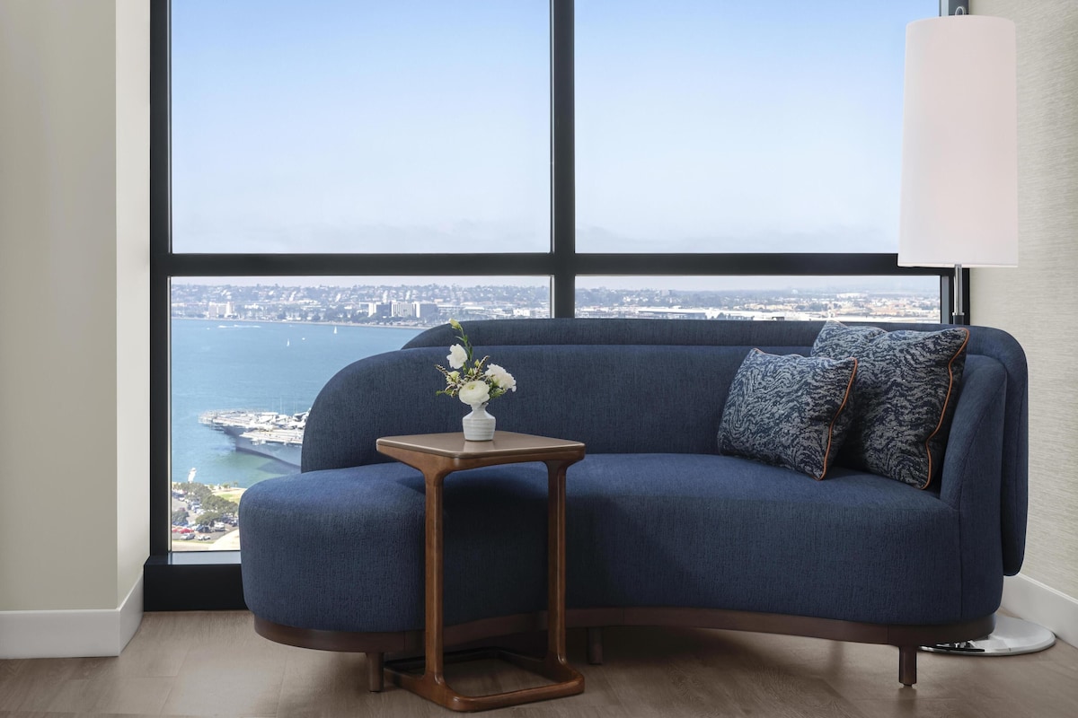 A room with a blue sofa, a small wooden side table with a vase of flowers, and a view of a body of water and distant buildings.