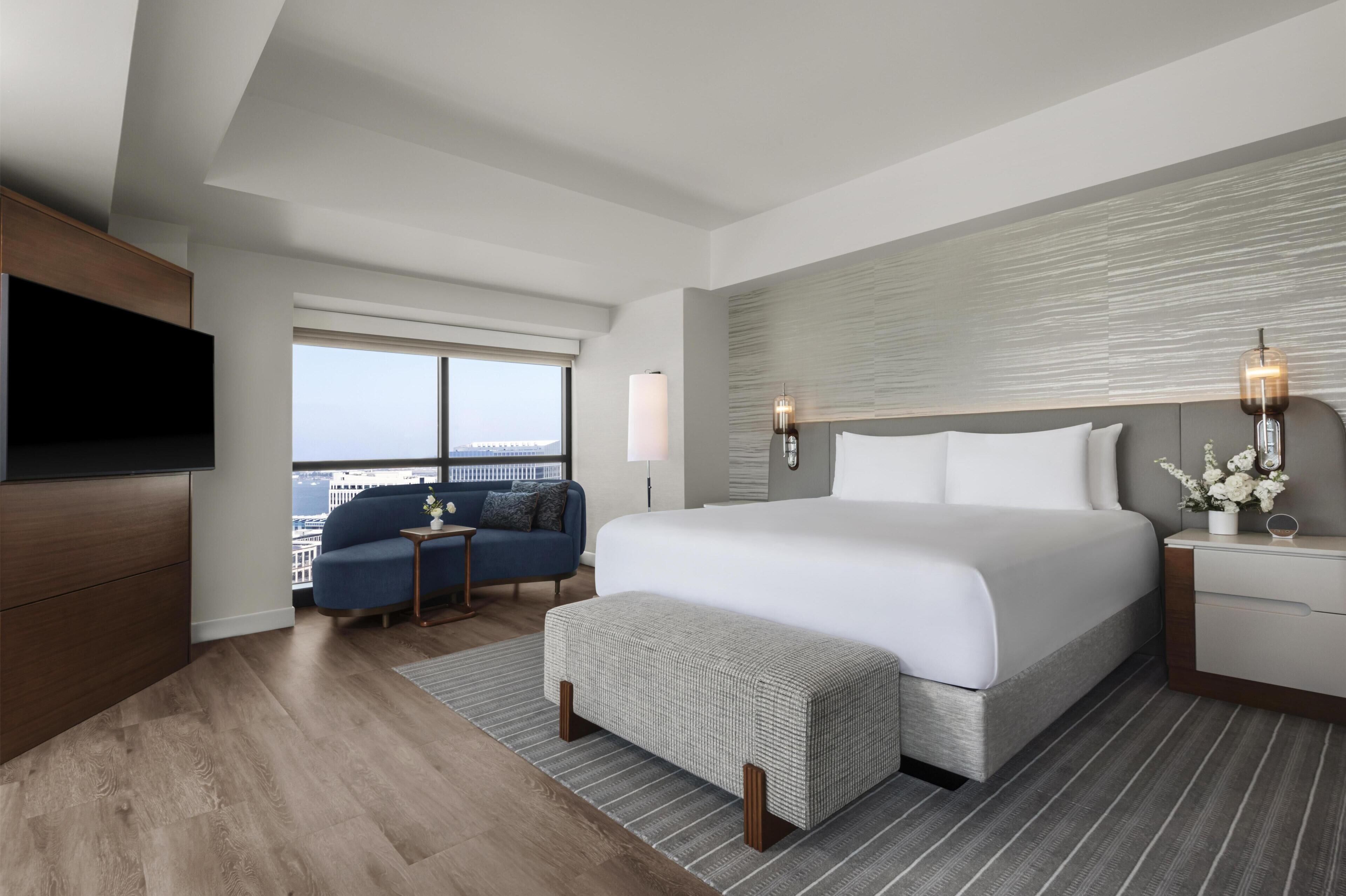 A modern hotel room with a large bed, a television, a sofa, and a view of the city.