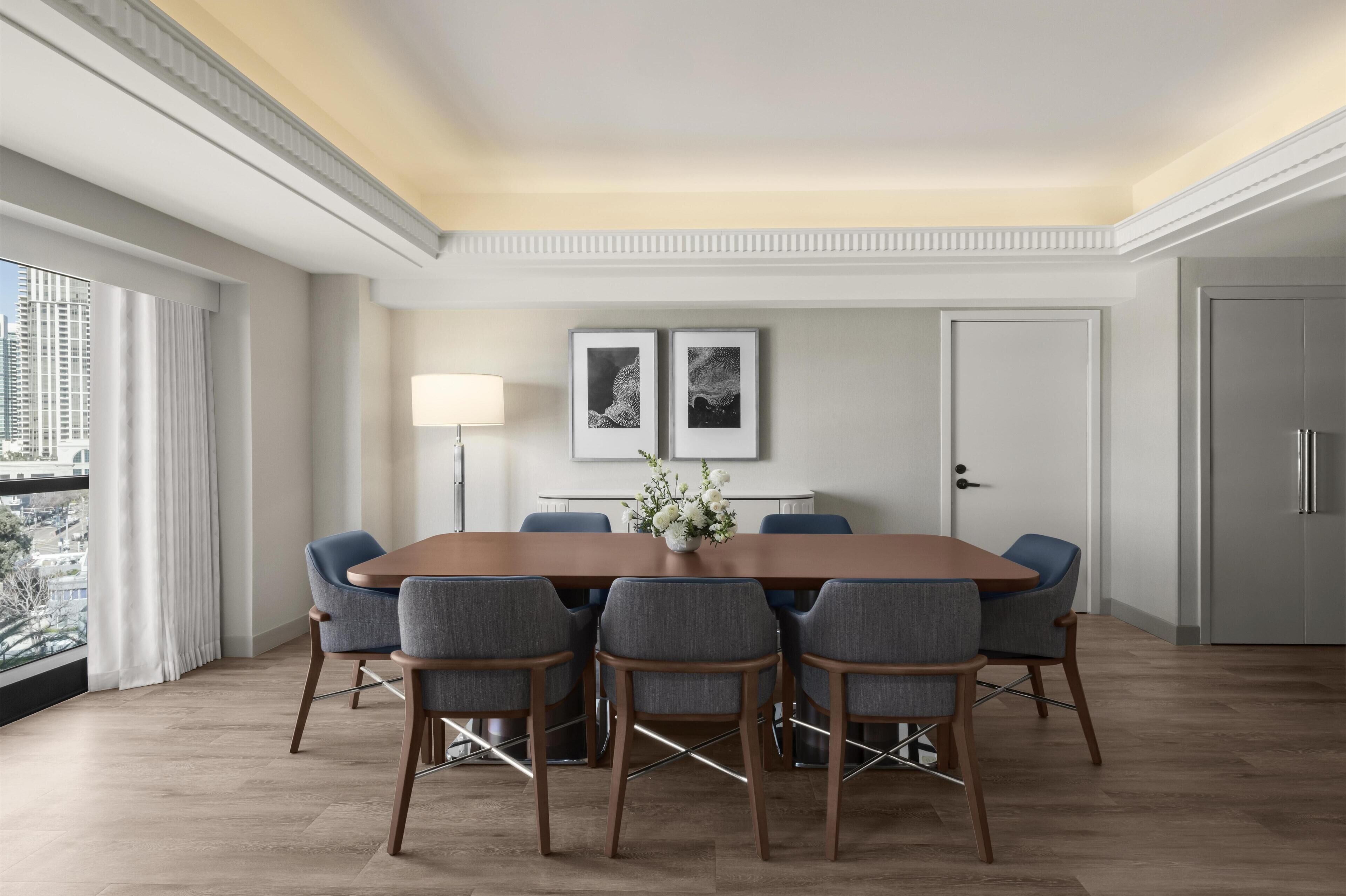 Un espace de salle à manger moderne avec une longue table, six chaises, un vase de fleurs, deux tableaux encadrés au mur et une vue sur la ville à travers de grandes fenêtres.