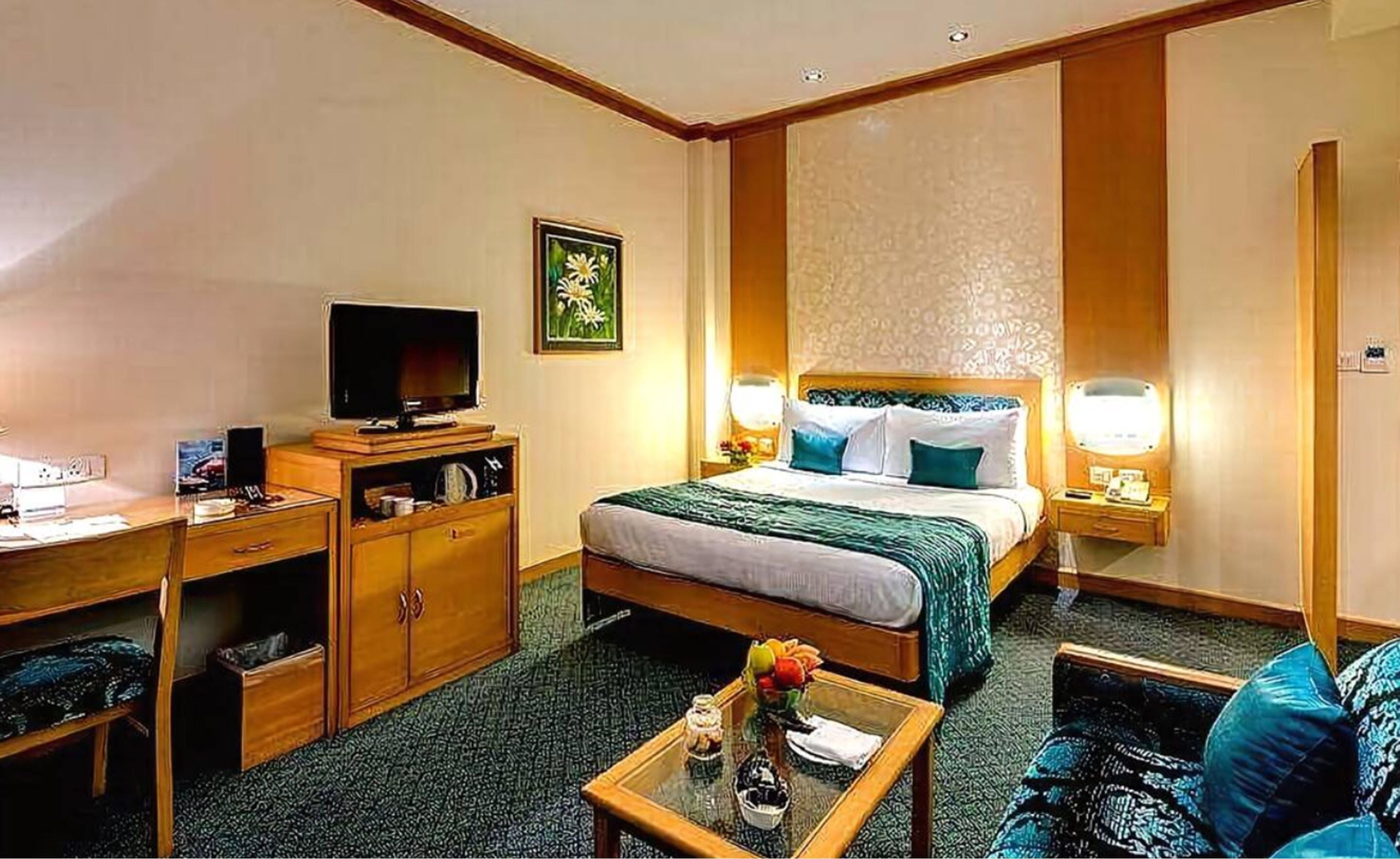 Habitación de hotel con cama, escritorio con televisor, cómoda, mesita de noche con lámpara y una pequeña mesa con un jarrón de flores.
