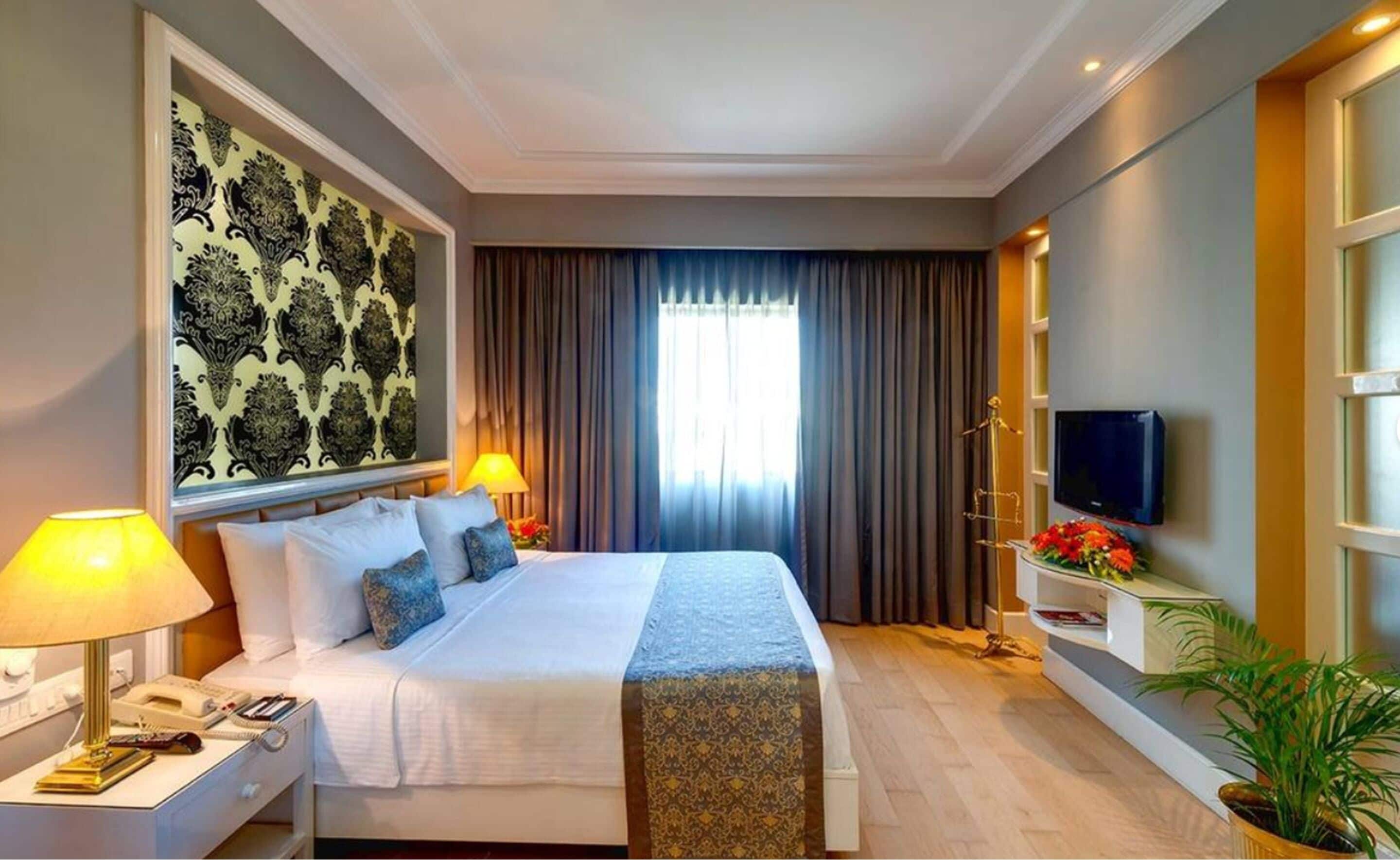 Habitación de hotel con cama, lámparas de noche, televisor, escritorio con teléfono y un ventanal con cortinas.