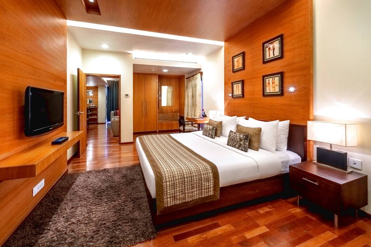 A hotel room with a wooden floor, a bed with a patterned bedspread, a television on a wooden stand, a nightstand with a lamp, and a wall with framed pictures.