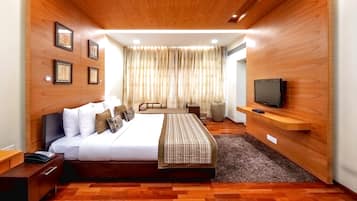 Una habitación de hotel moderna con techo de madera, una cama grande con ropa blanca, un televisor sobre una estantería de madera y un ventanal con cortinas.