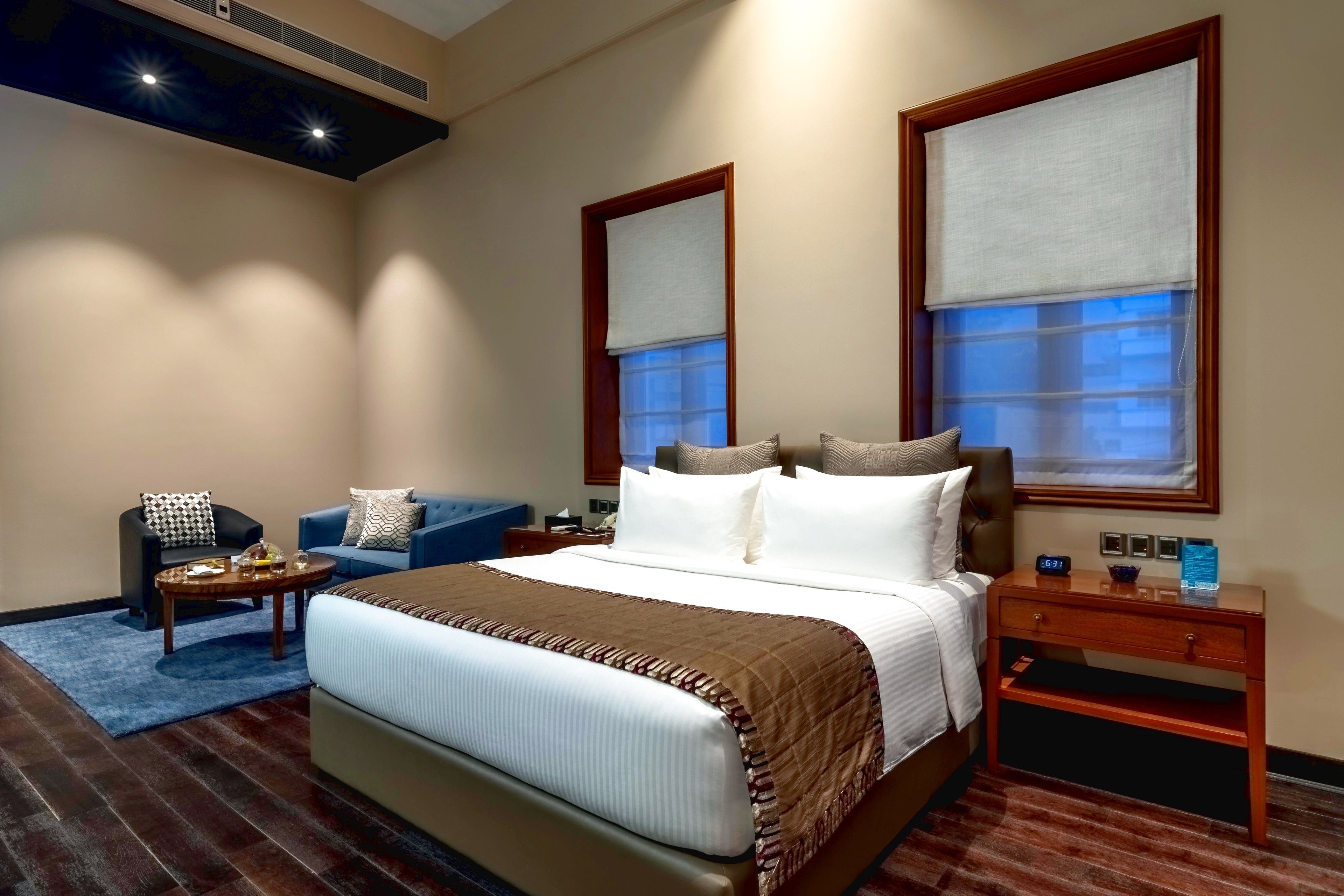 Habitación de hotel con una cama grande, una zona de estar con sofá y sillón, una mesita de noche de madera y una ventana con vistas.