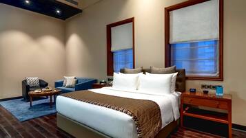Habitación de hotel con una cama grande, una zona de estar con sofá y sillón, una mesita de noche de madera y una ventana con vistas.