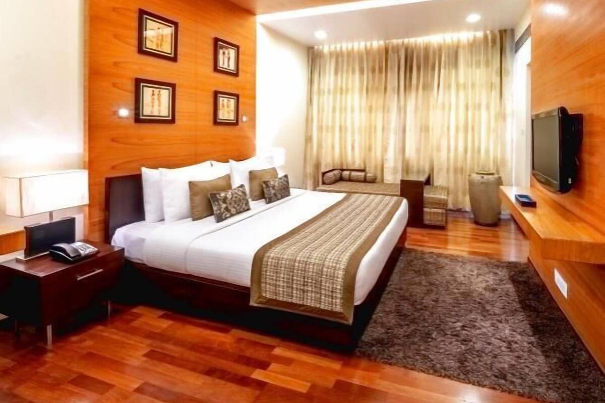 A hotel room with a wooden ceiling, a bed with white bedding, a TV on the wall, and a desk with a lamp.