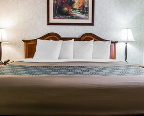 A neatly made bed with a patterned bedspread and a wooden headboard. There are two bedside lamps and a painting on the wall above the bed. - Econo Lodge & Suites Grand Rapids Airport (Grand Rapids)