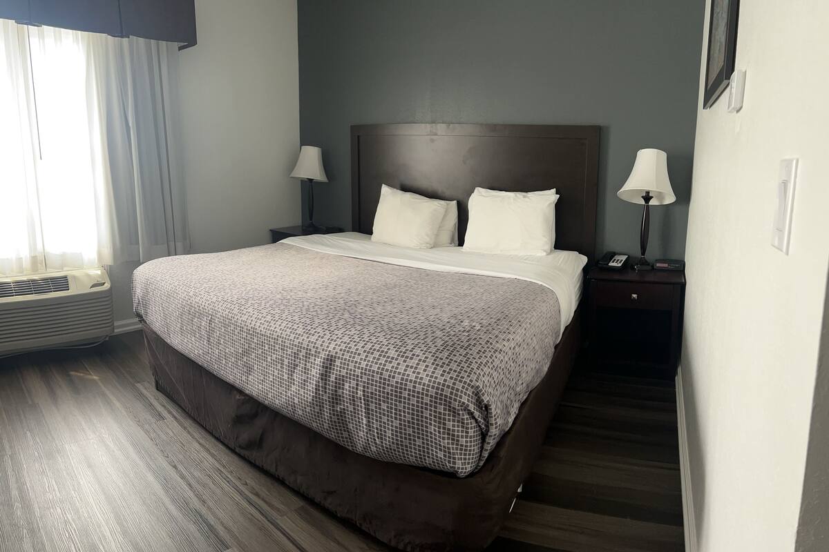 A hotel room with a large bed, two bedside tables with lamps, a window with curtains, and a wall-mounted picture.
