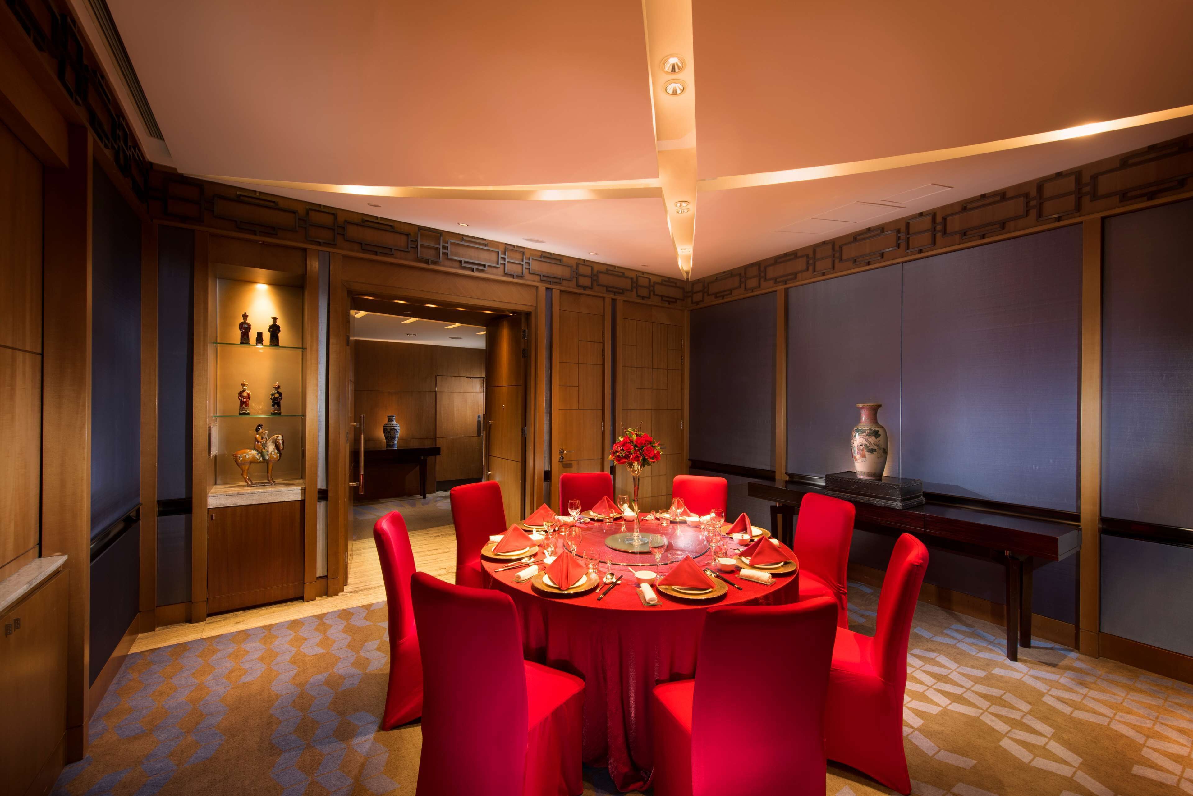 Un comedor con una mesa redonda dispuesta para seis personas, rodeada de sillas rojas. Hay un jarrón con flores sobre la mesa, y la habitación tiene paneles de madera y una alfombra estampada.