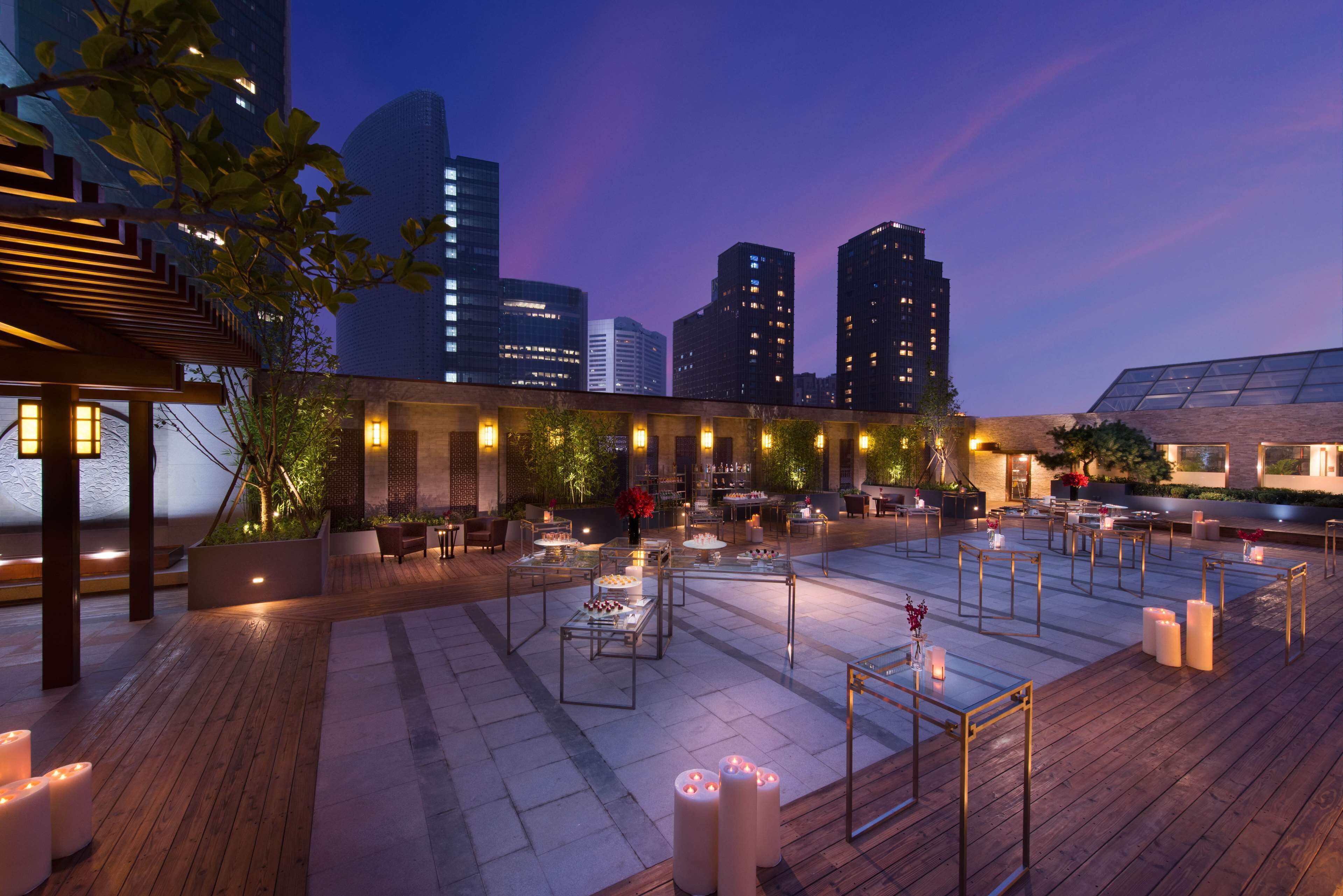 An outdoor dining area with tables and chairs, surrounded by plants and candles, with a city skyline in the background.