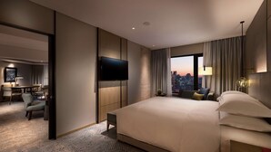 Une chambre d’hôtel moderne dotée d’un grand lit, d’un téléviseur à écran plat fixé au mur, offrant une vue sur la ville depuis la fenêtre, et d’un coin salon séparé avec une table et des chaises.