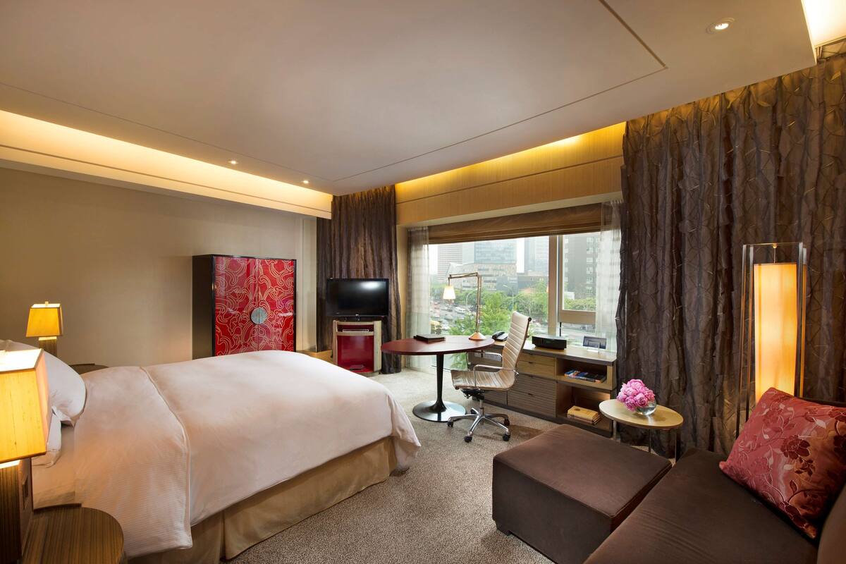 Habitación de hotel con cama, escritorio, silla, televisor, ventana con vista a la ciudad y una mesita con un jarrón de flores.