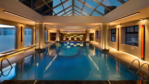 A modern indoor swimming pool with glass walls, a skylight, and lounge chairs.