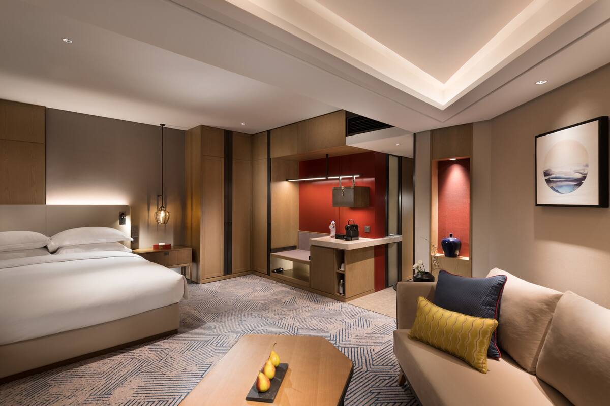 Una habitación de hotel moderna con una cama grande, un sofá, una pequeña mesa con fruta, un escritorio y un espejo.