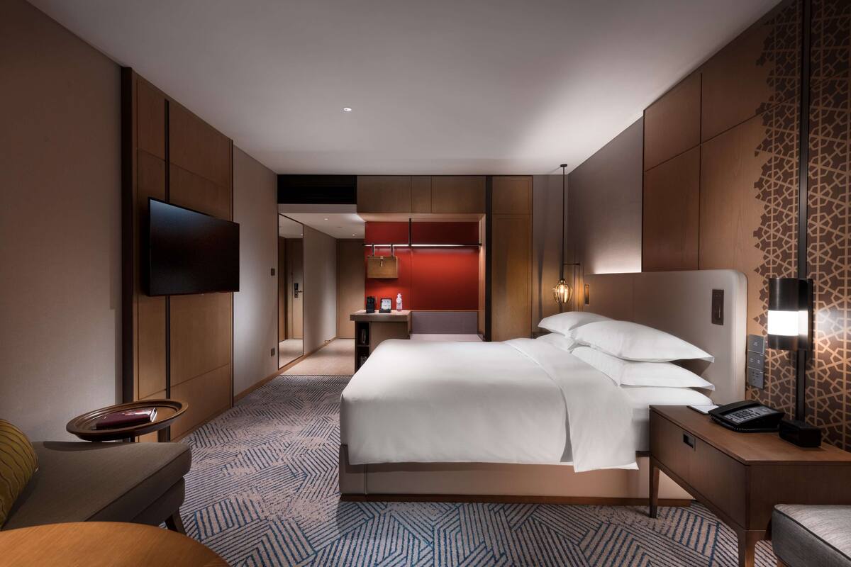 Una habitación de hotel moderna con una cama grande, un televisor montado en la pared, un escritorio con lámpara y una pequeña zona de estar.