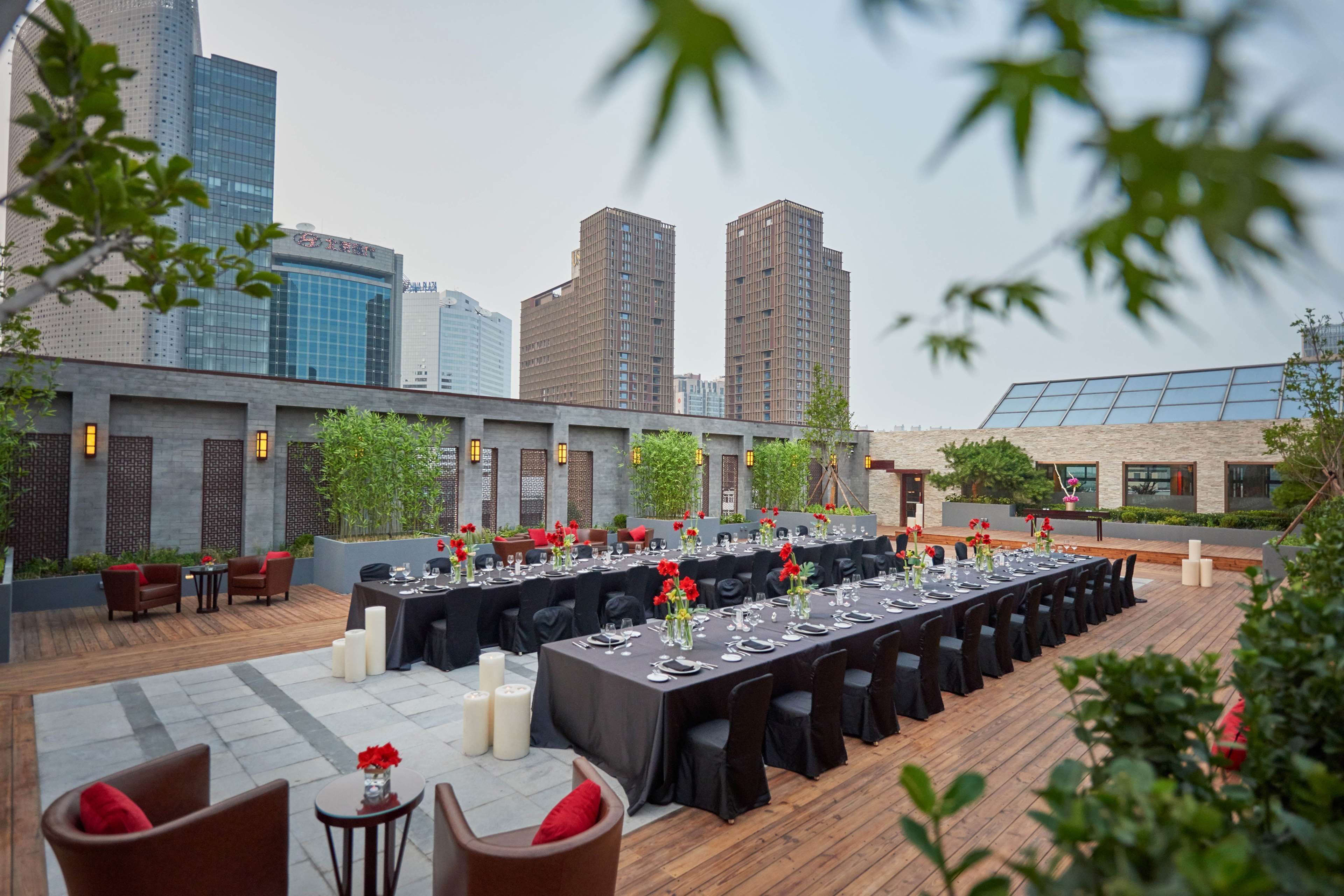 An outdoor dining area with tables set for a meal, surrounded by city buildings and greenery.