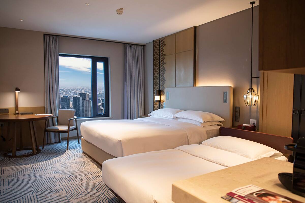 Habitación de hotel con dos camas, un escritorio, una silla, vista a la ciudad y un ventanal grande.