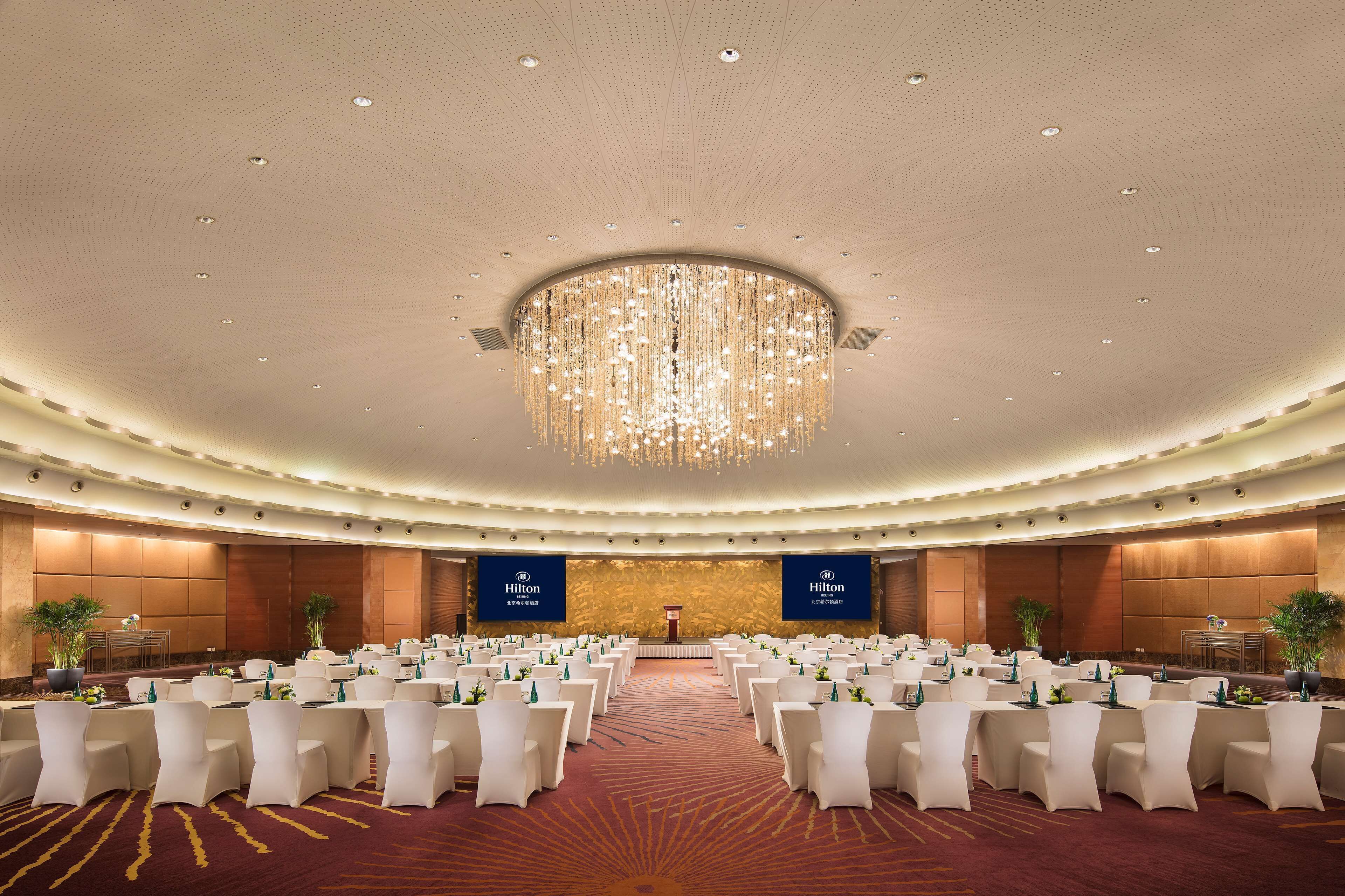 A large, well-lit conference room with rows of white chairs, a central stage, and two Hilton banners.