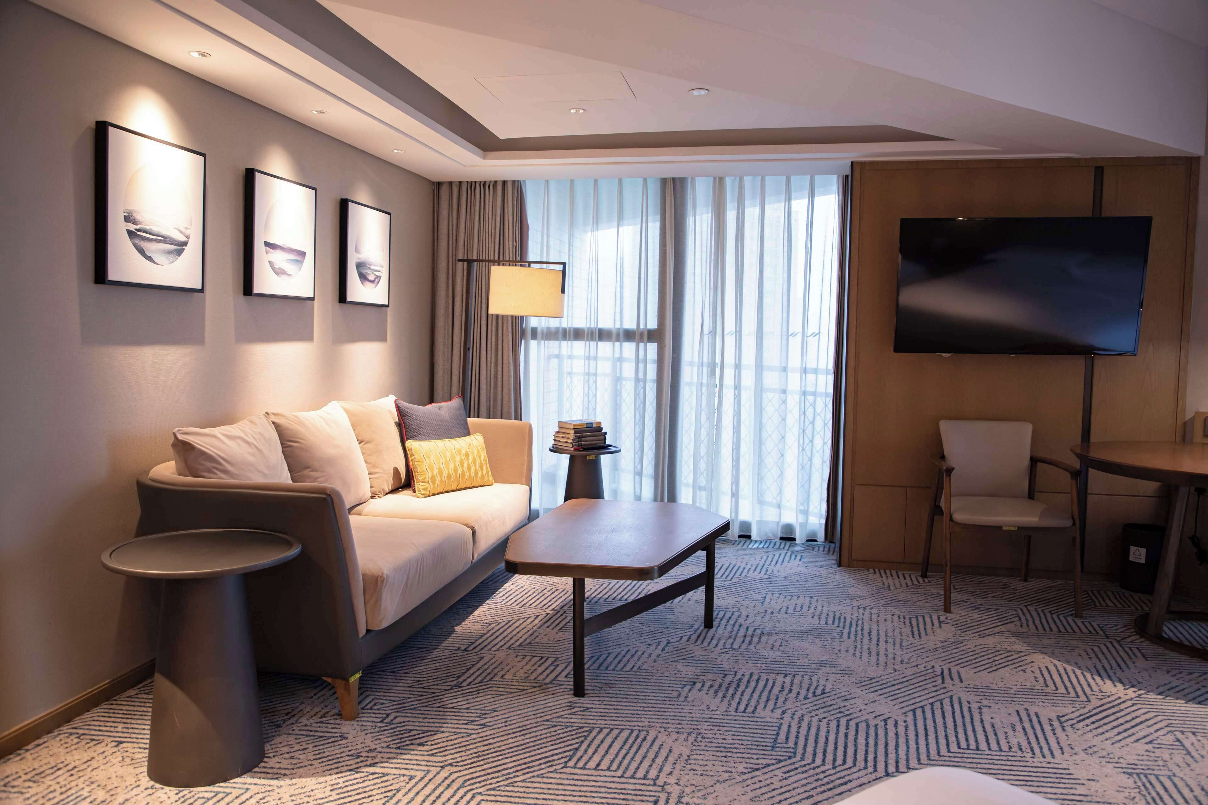 A modern hotel room with a sofa, a coffee table, a TV, and a dining area.