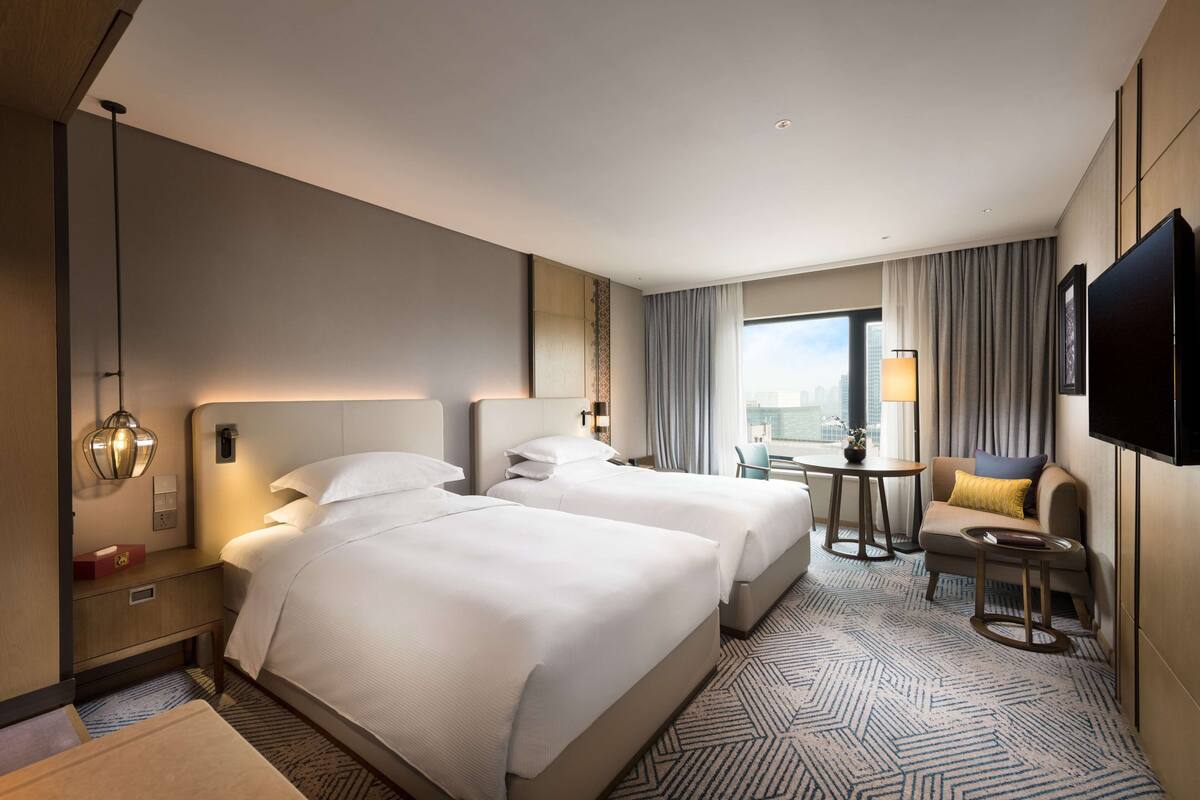 Habitación de hotel con dos camas, un escritorio, una silla, un televisor y un ventanal con vistas a la ciudad.