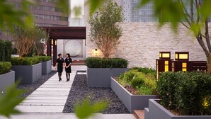 Une entrée d’hôtel moderne, avec un chemin menant à la réception, entourée de jardins aménagés et d’un grand immeuble en arrière-plan.
