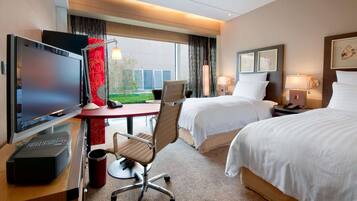 A hotel room with a desk, TV, two beds, and a view of a building outside.