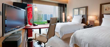A hotel room with a desk, TV, two beds, and a view of a building outside.