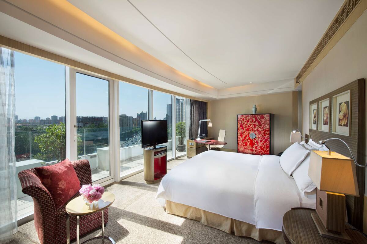 Habitación de hotel con una cama grande, televisión, escritorio, silla y balcón con vista a la ciudad.
