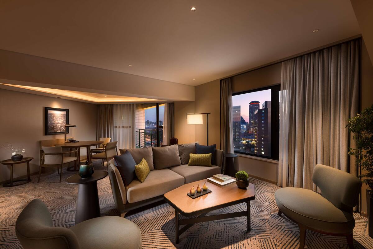 Habitación de hotel moderna con sofá, sillones, una mesita de centro y vistas a la ciudad.