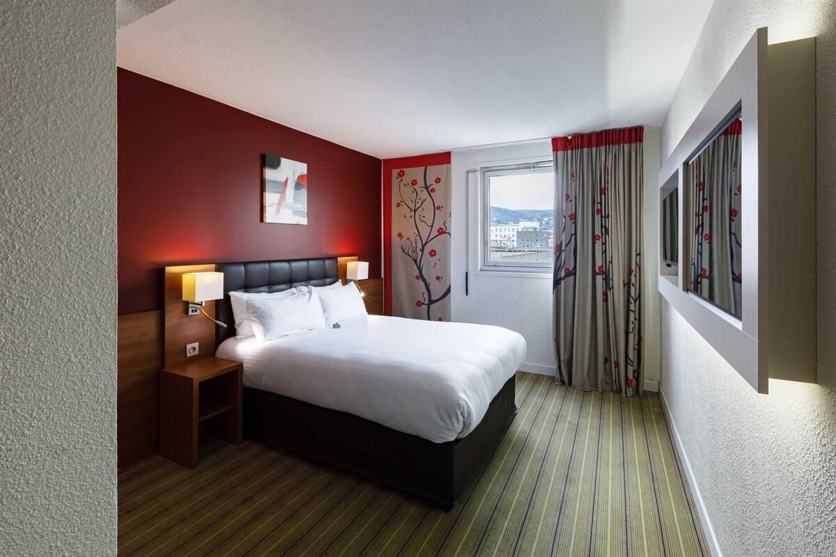 A hotel room with a bed, nightstand, wall art, window with curtains, and a view of buildings.