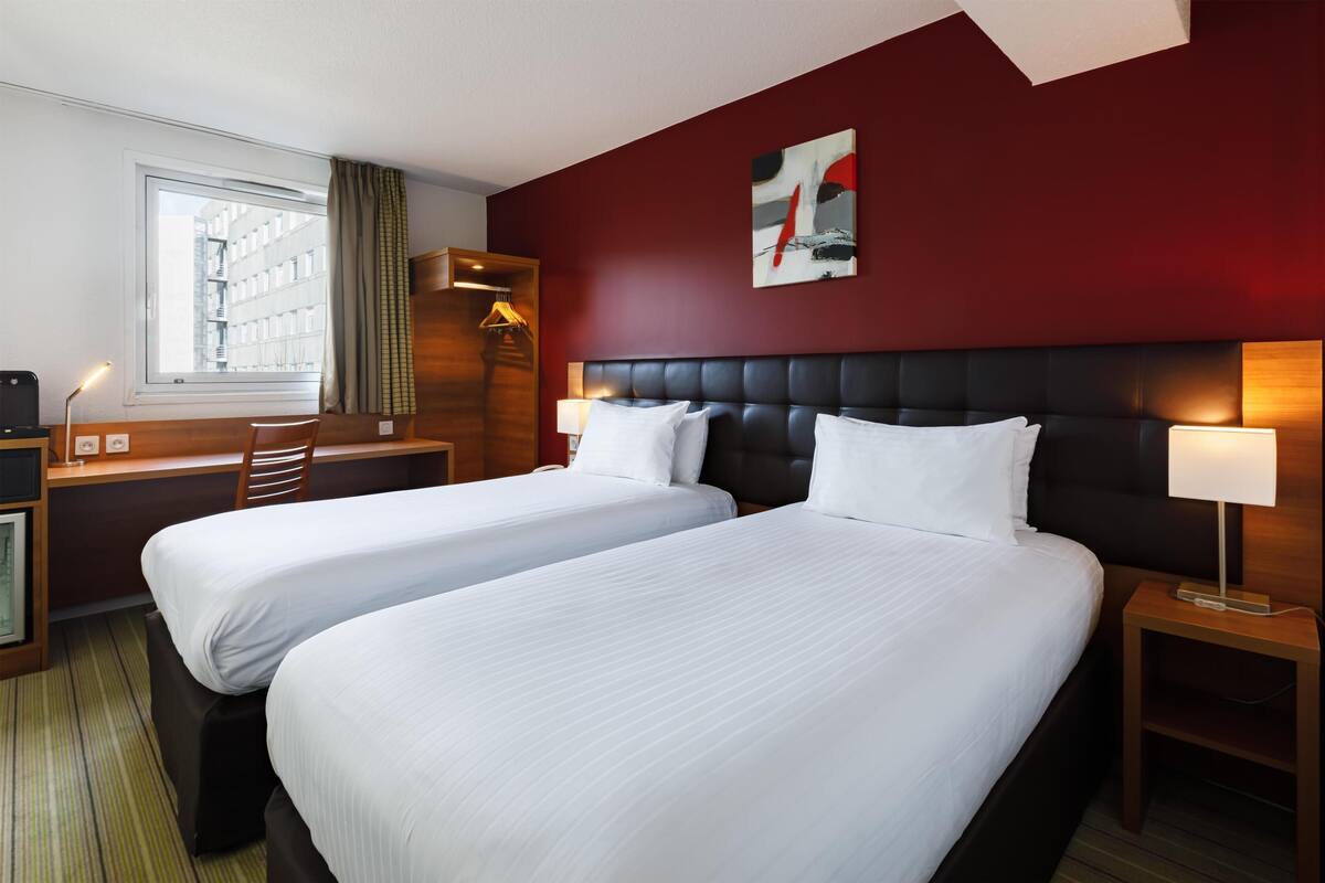 A hotel room with two beds, a desk, a chair, a lamp, and a window with a view of buildings.