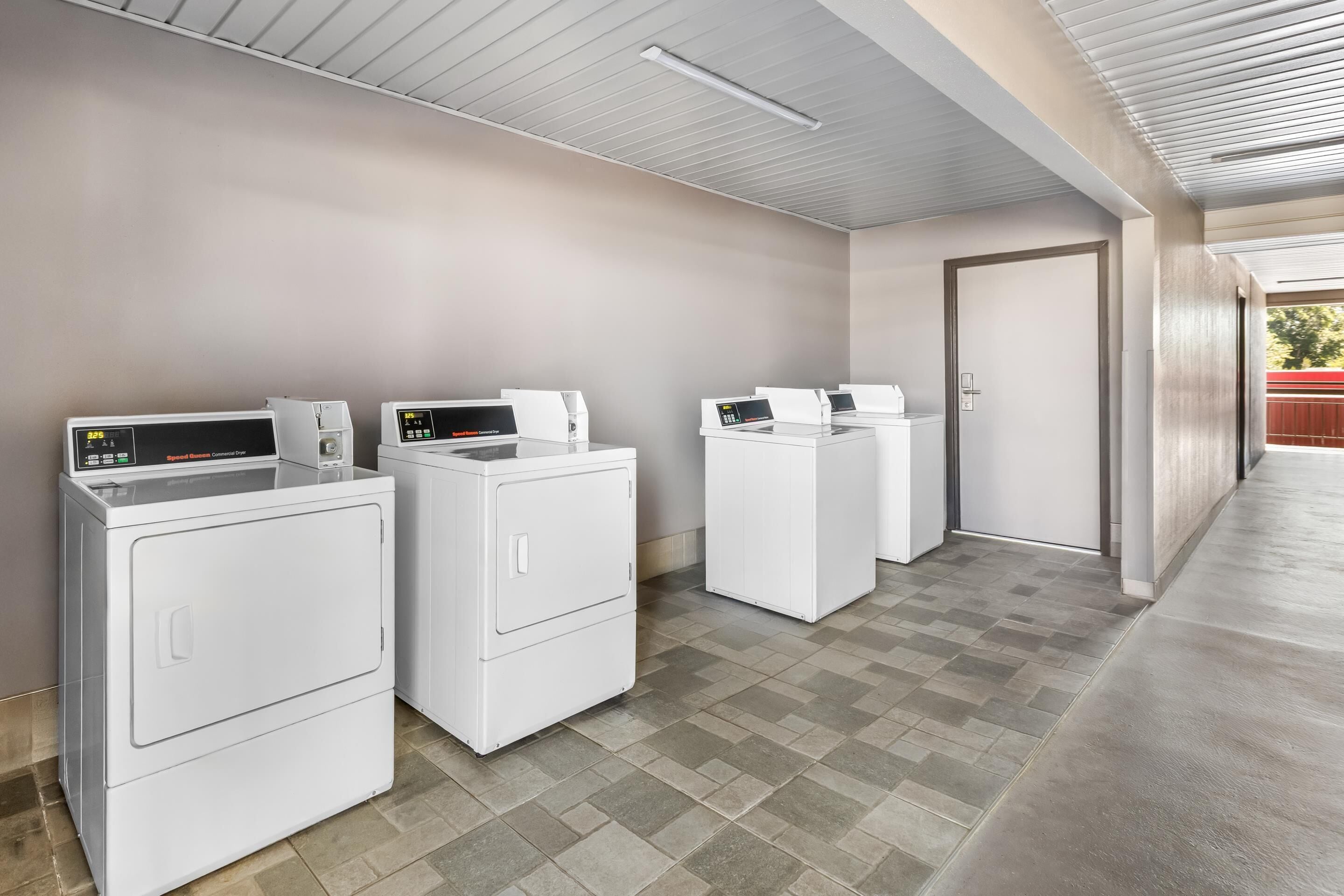 A laundry area with three white front-loading washers and dryers, a door to the right, and a tiled floor.
