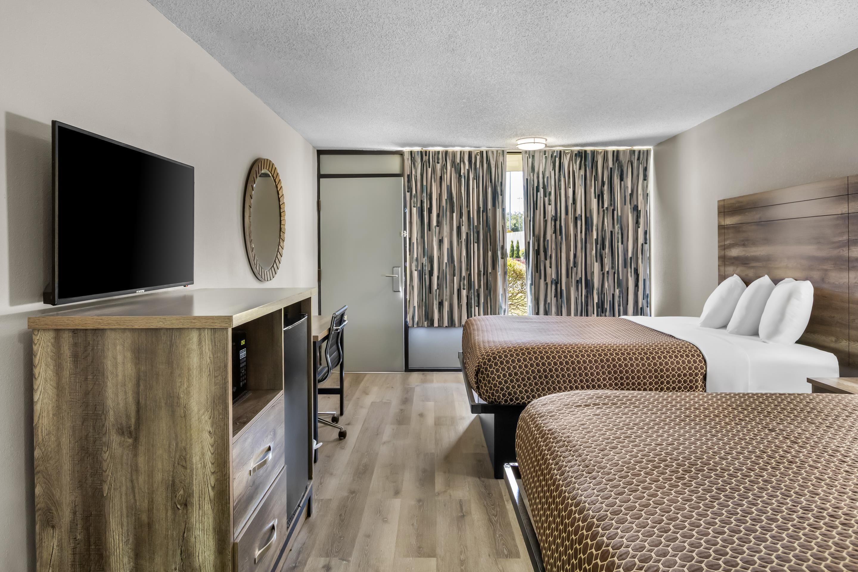 A hotel room with a TV, a desk, a chair, a bed with a patterned bedspread, and a wooden headboard.