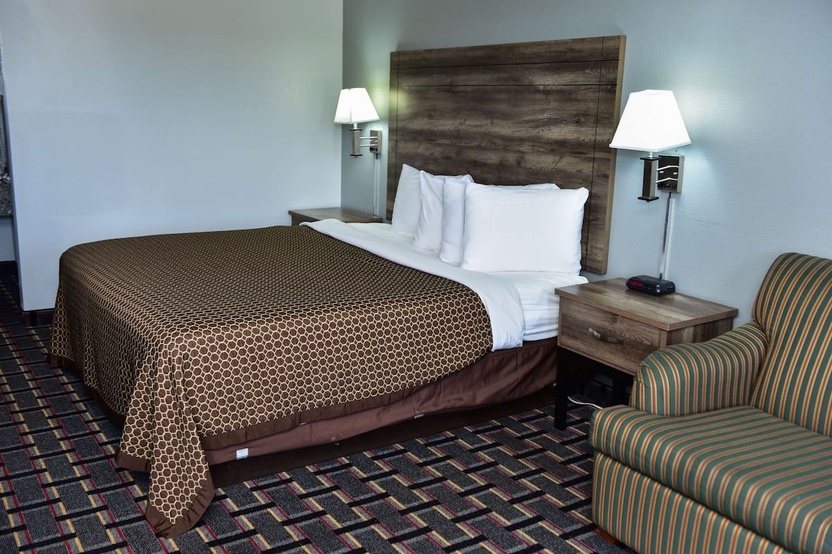 A hotel room with a large bed, two bedside lamps, a wooden headboard, a nightstand, a striped armchair, and a patterned carpet.
