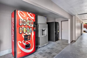 A vending machine with various beverage options and a refrigerator in a hallway.