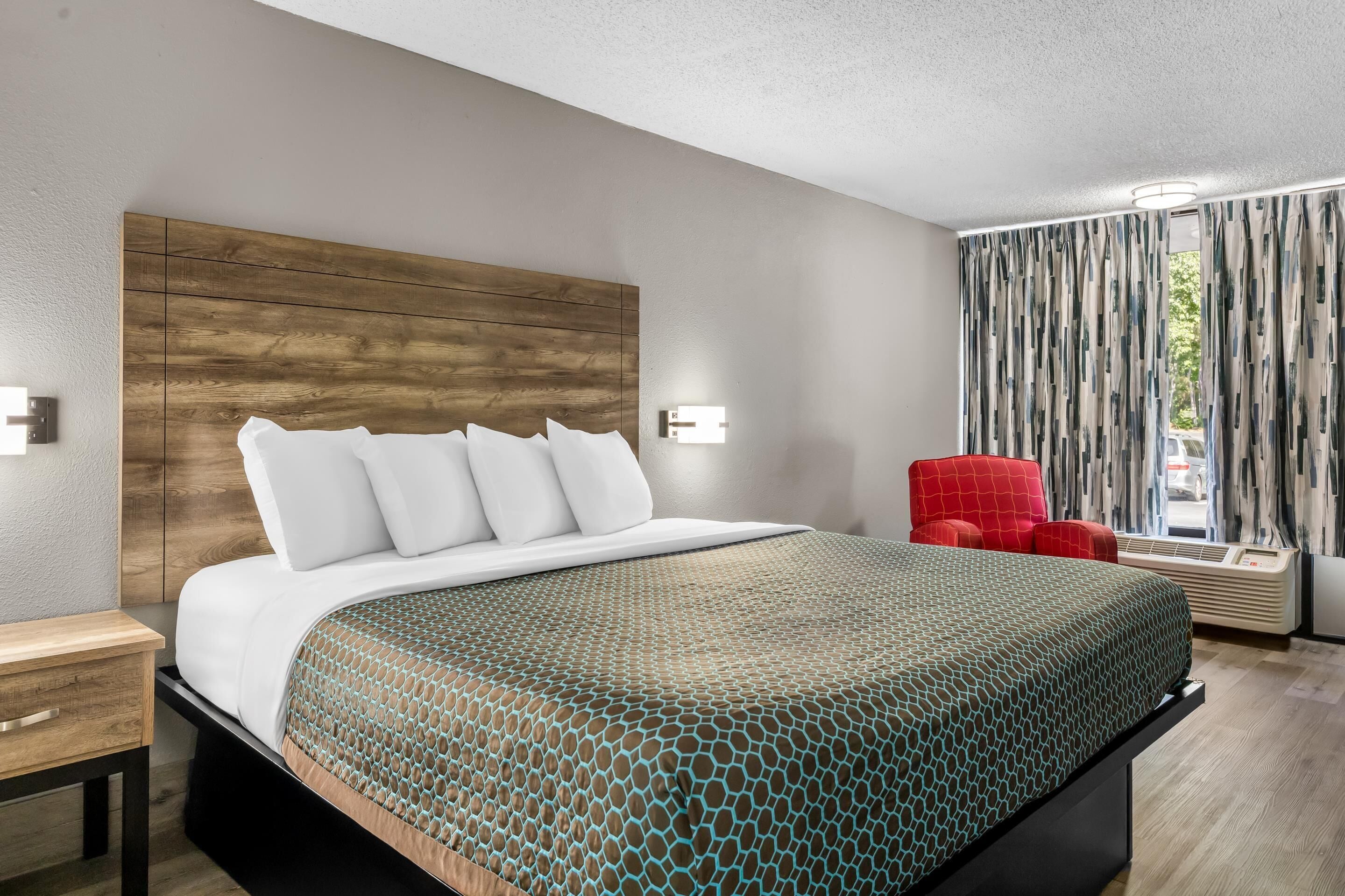 Ein modernes Hotelzimmer mit einem hölzernen Kopfteil, einem großen Bett mit gemusterten Betttextilien, einem roten Sessel und Blick ins Freie durch Vorhänge.