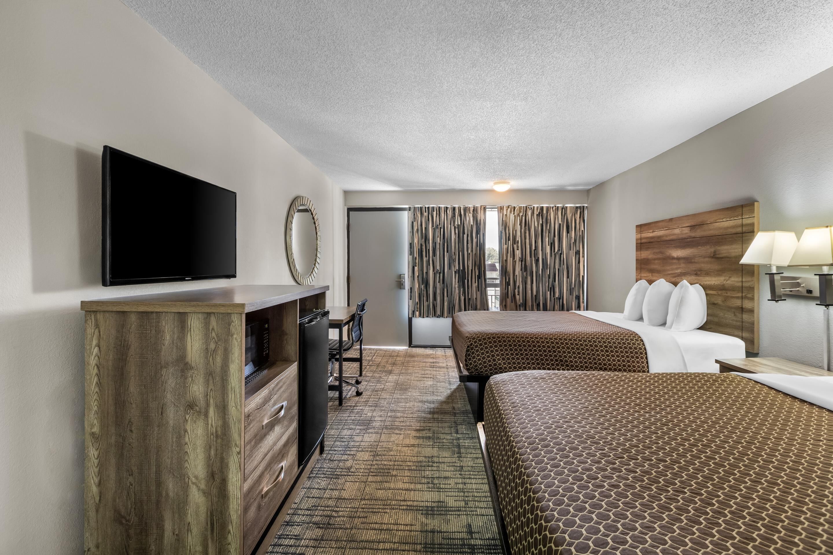 A hotel room with a flat-screen TV, wooden furniture, a desk, a chair, two beds with patterned bedding, a mirror, curtains, and a lamp.