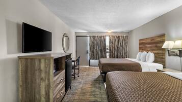A hotel room with a flat-screen TV, wooden furniture, a desk, a chair, two beds with patterned bedding, a mirror, curtains, and a lamp.