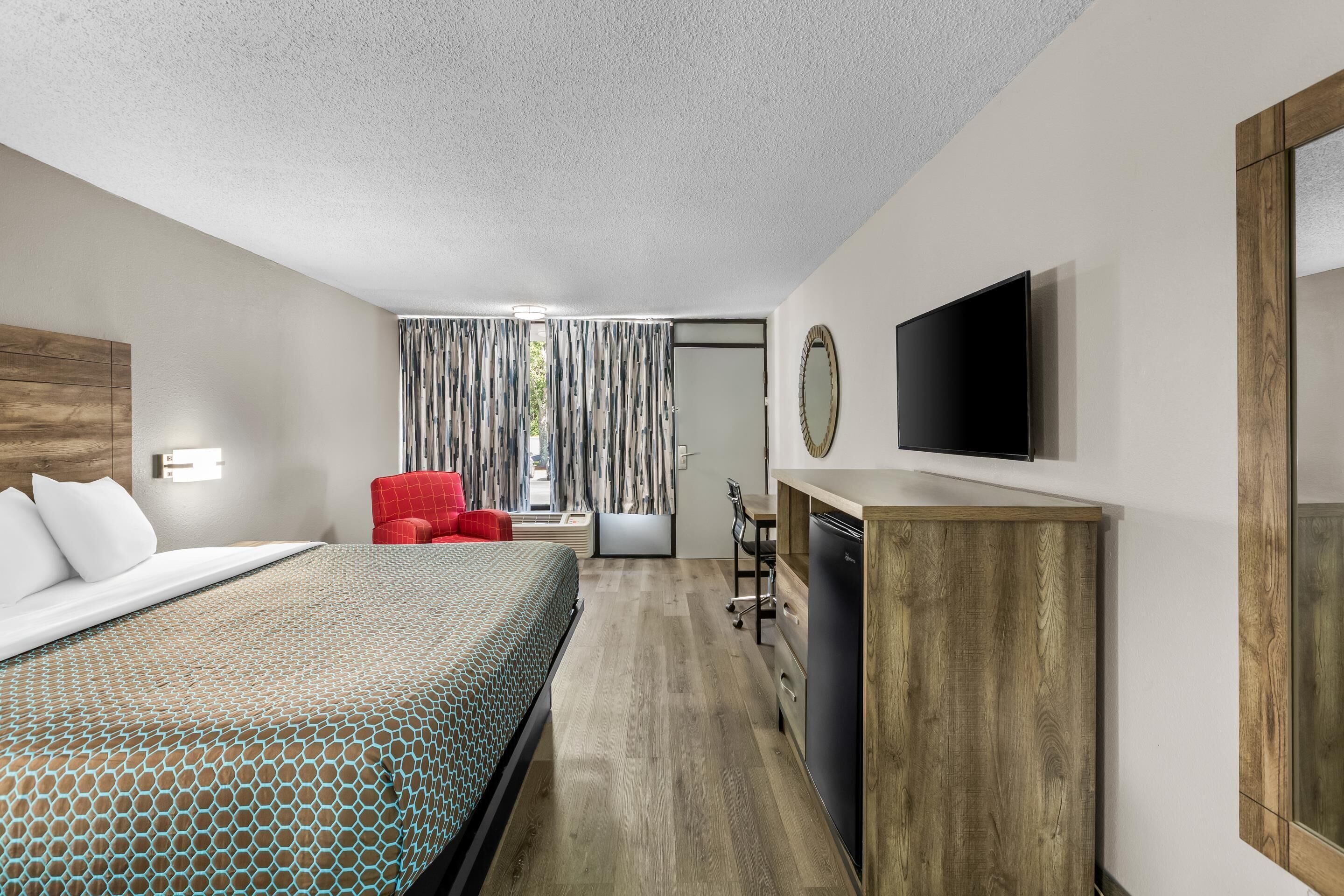 A hotel room with a bed, a red chair, a desk with a flat-screen TV, and a mirror.