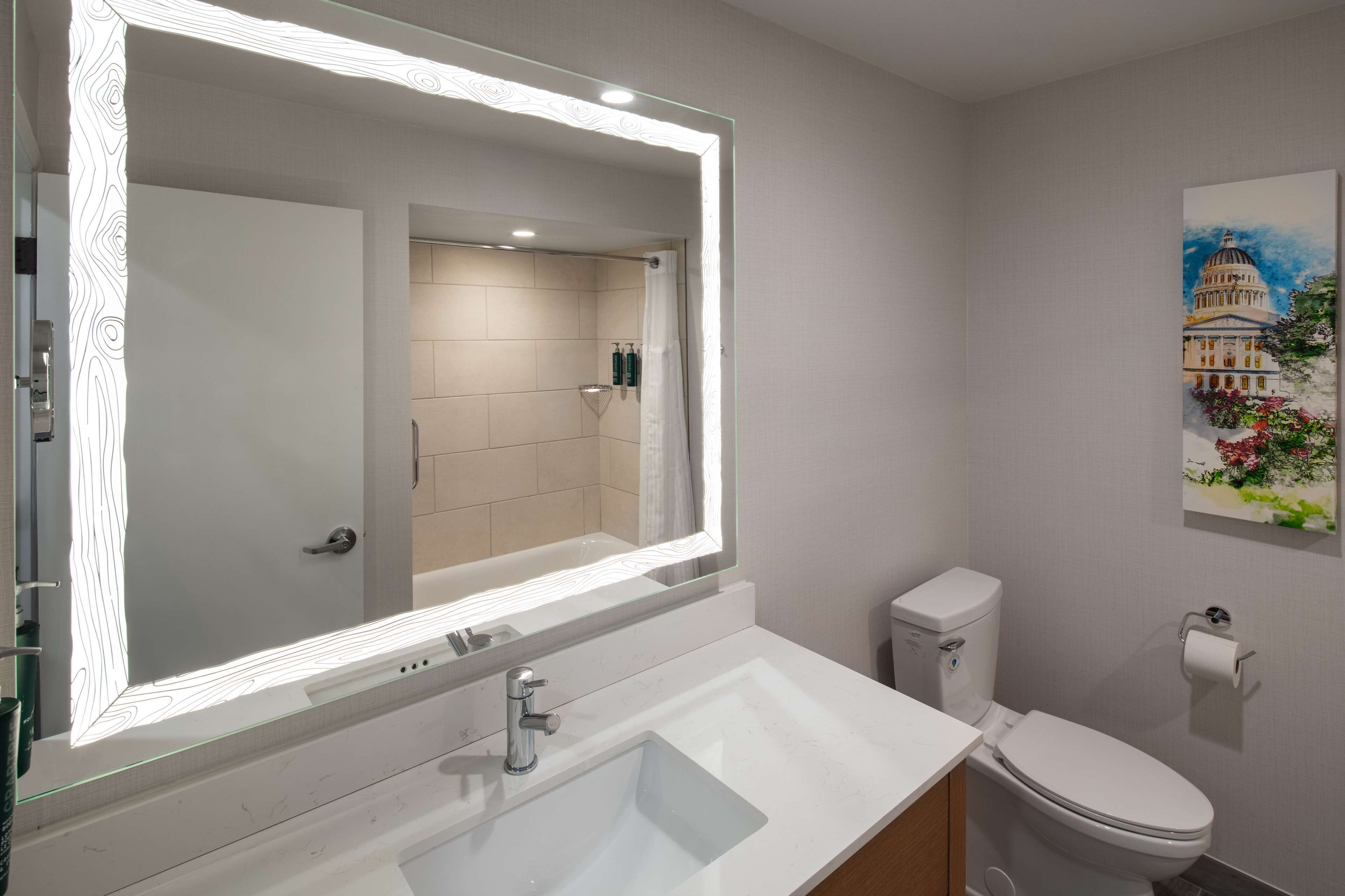 A bathroom with a large mirror, a sink, a toilet, and a painting of a building on the wall.