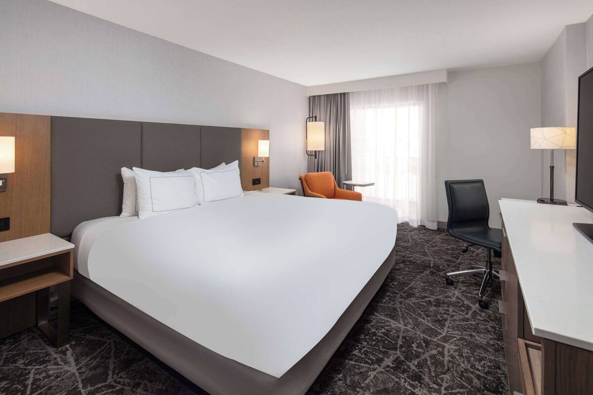 A hotel room with a large bed, a desk, an orange chair, and a TV.