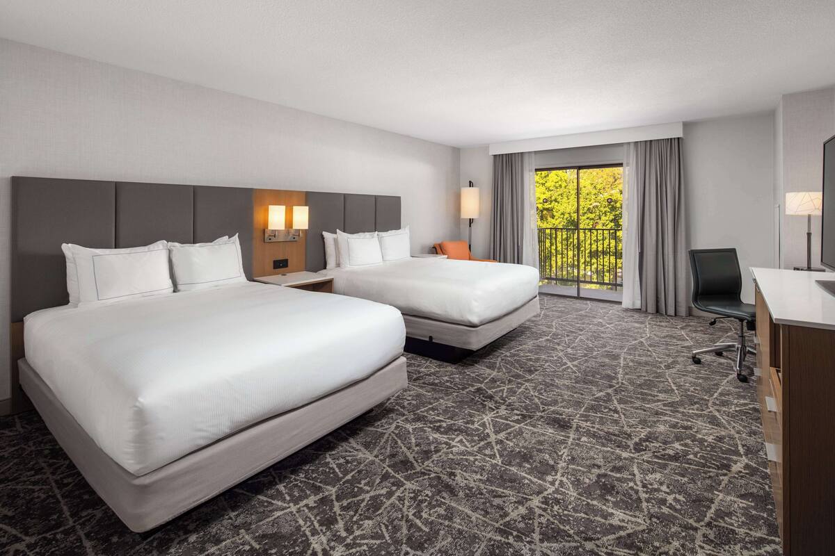 A hotel room with two beds, a desk, and a view of greenery through the window.