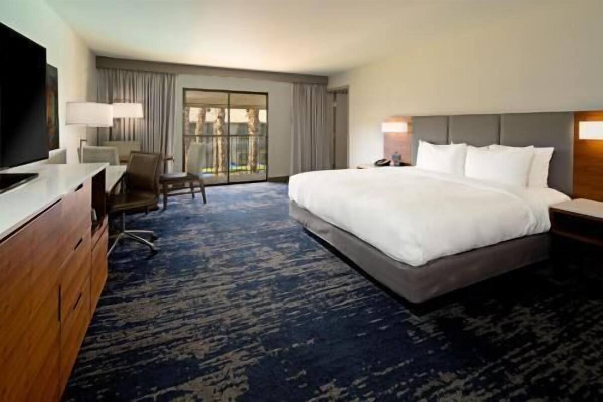 A hotel room with a large bed, a desk, a chair, a television, and a view of the outdoors through a sliding door.