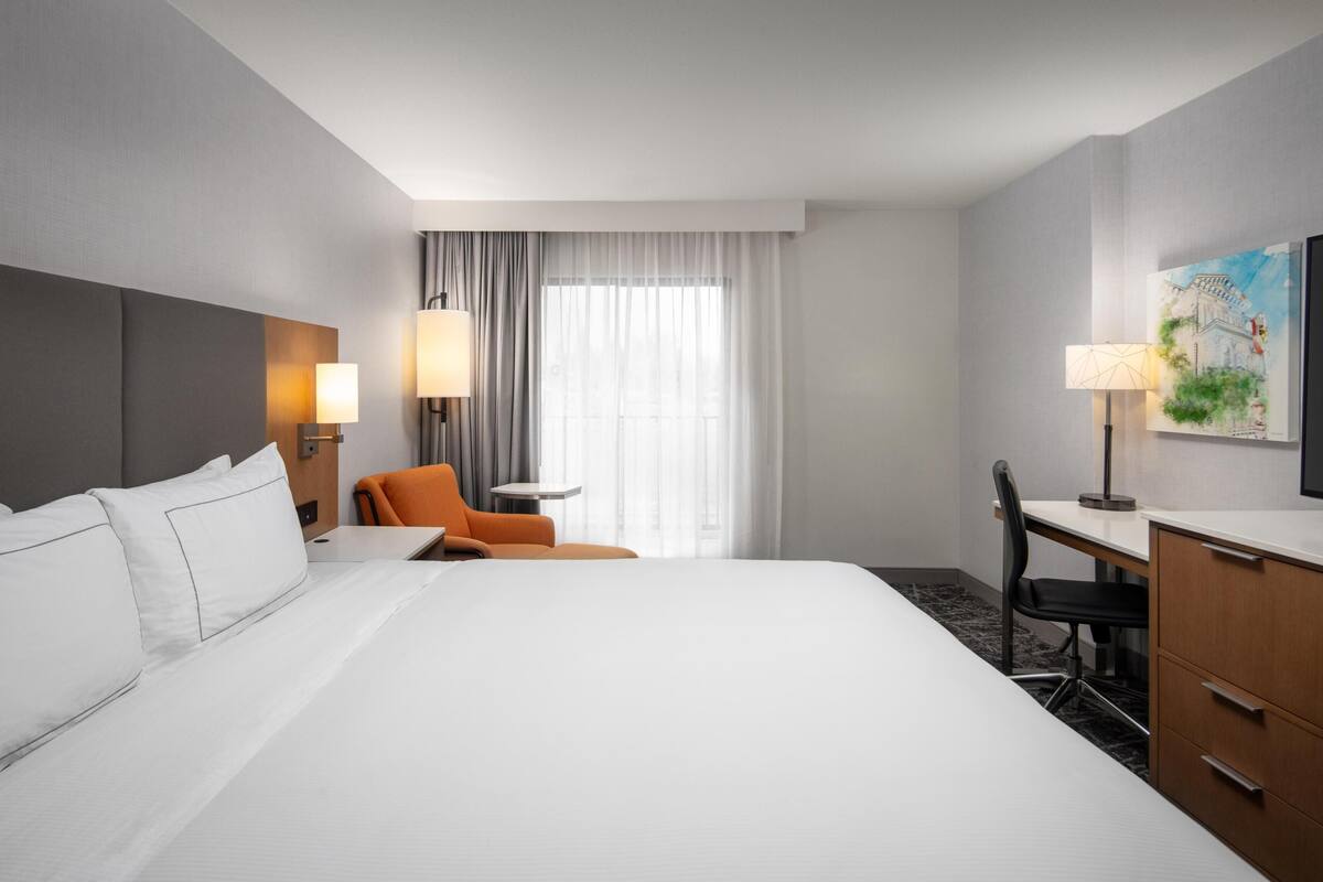 A hotel room with a large bed, a desk, an orange chair, a TV, and a painting on the wall.