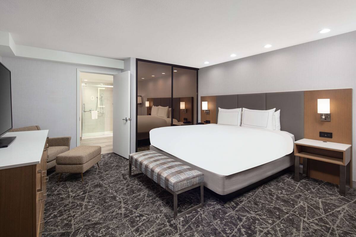 A modern hotel room with a large bed, a desk, a chair, a bench, and a TV.