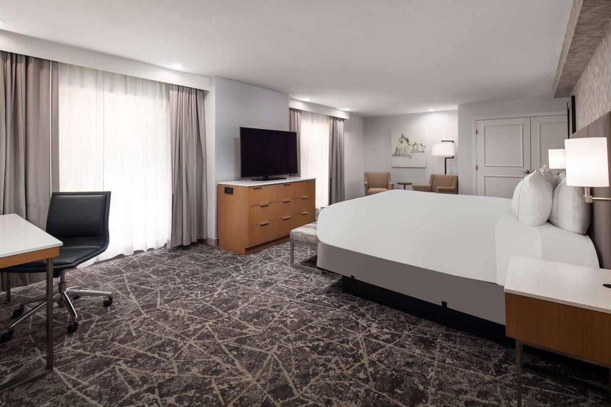 A hotel room with a large bed, a desk with a chair, a TV on a wooden dresser, and a lamp on a bedside table.
