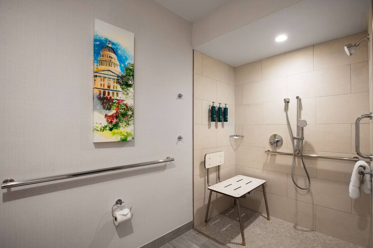 A modern bathroom with a handrail, a small chair, a shower with a handheld showerhead, and a painting of a building on the wall.