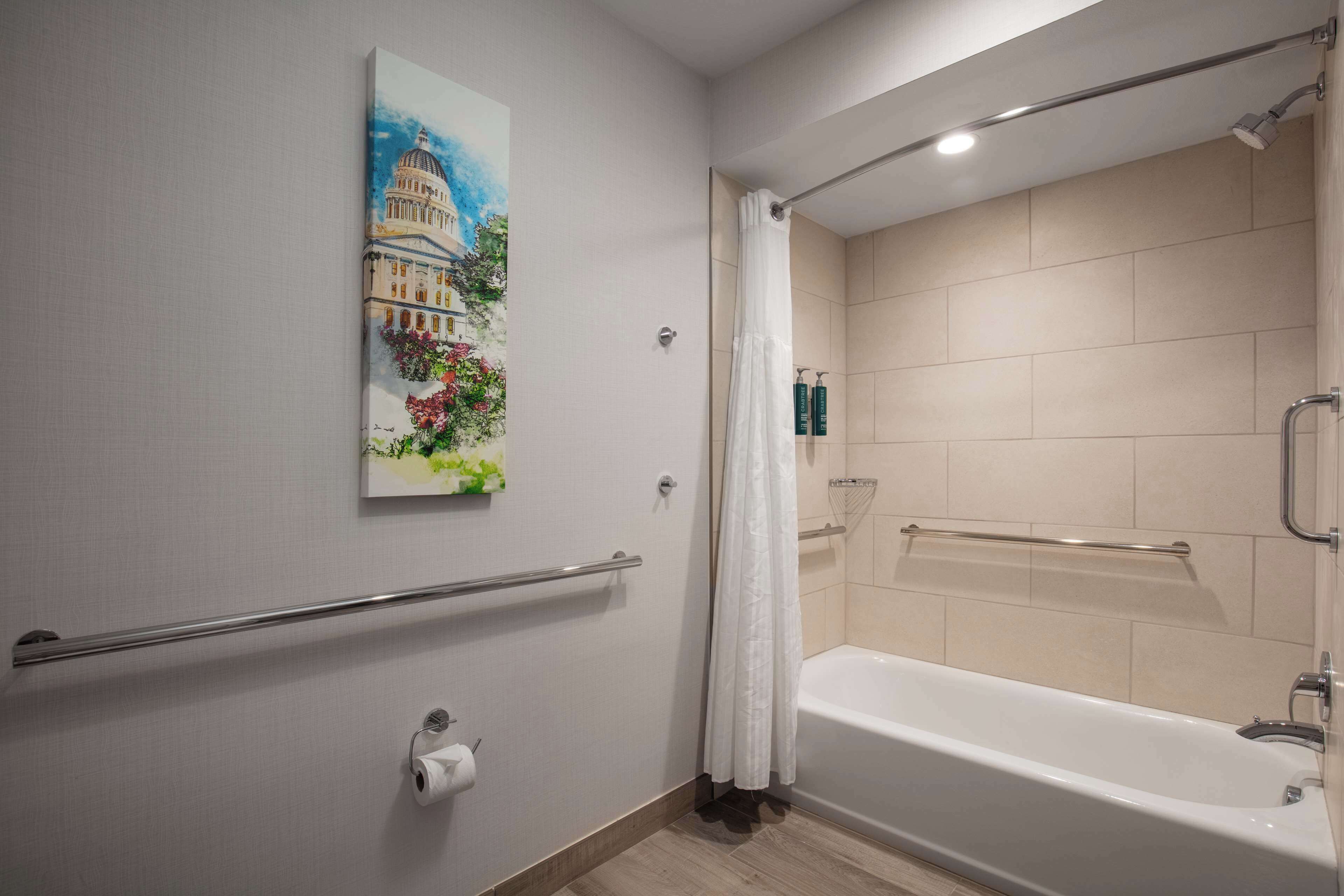 Une salle de bain moderne avec une baignoire, une douche dotée d’une porte en verre, une barre d’appui et un tableau représentant un bâtiment accroché au mur.