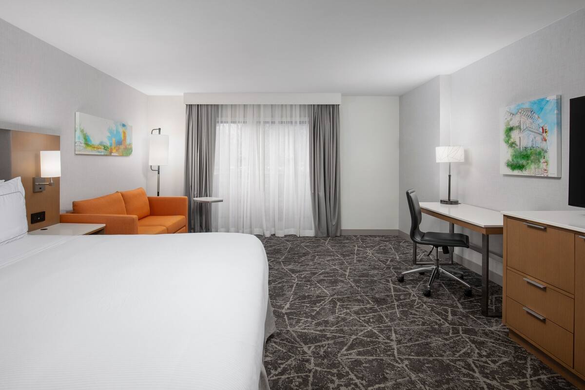 A hotel room with a large bed, an orange sofa, a desk with a chair, a television, and artwork on the walls.