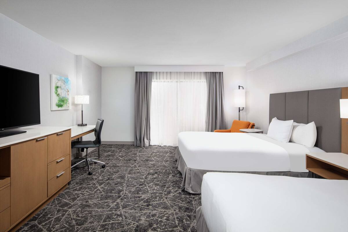 A hotel room with a TV, desk, two beds, and a large window with curtains.